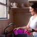 A young woman in glasses washes dishes by a window in a vintage-style kitchen.